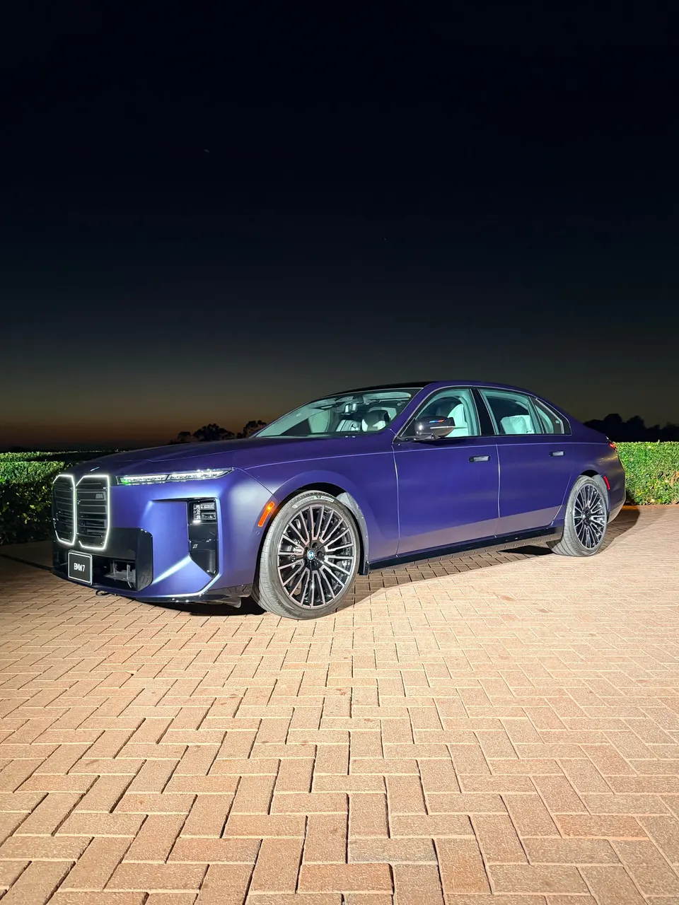 The 2027 BMW M760 xDrive's muted blue paint and illuminated grille stand out against the twilight sky.
